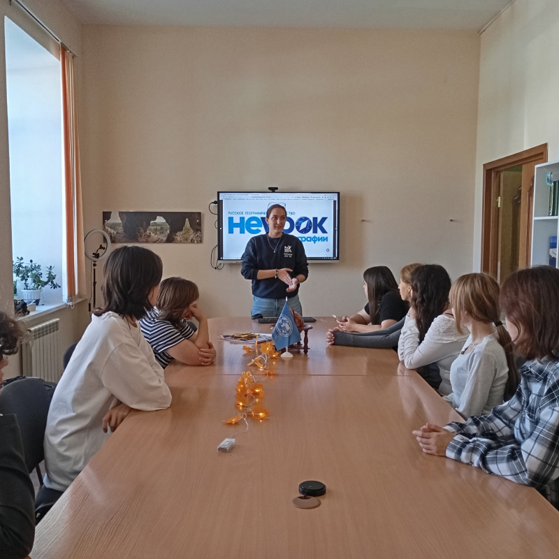 Девушка стоит у экрана с логотипом «НЕурок географии» и обращается к участникам