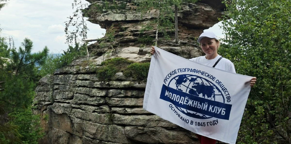 Руководитель клуба с флагом РГО на фоне скалы в Каменном городе