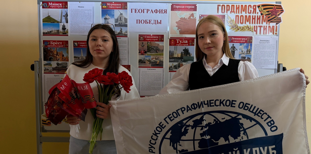 Участницы мероприятия с флагом Молодёжного клуба РГО и красными гвоздиками у стенда «География Победы»