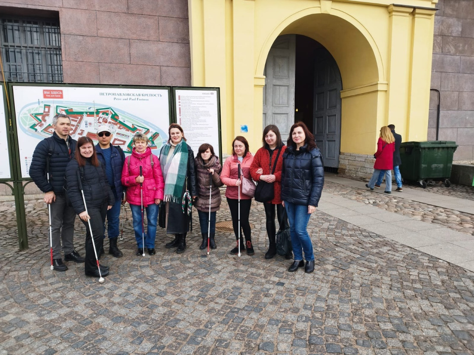 Инклюзивная группа незрячих петербуржцев и волонтёров на фоне стенда со схемой Петропавловской крепости