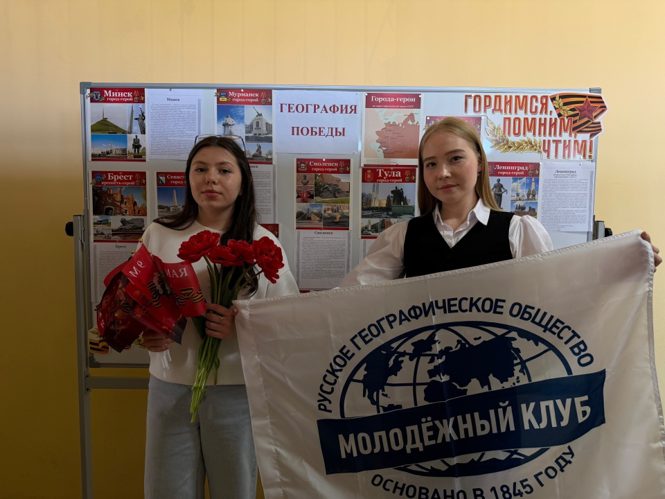 Участницы мероприятия с флагом Молодёжного клуба РГО и красными гвоздиками у стенда «География Победы»